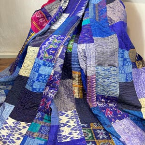 May include: A patchwork quilt in shades of blue, purple, and teal. The quilt is made of various fabric pieces sewn together, creating a textured surface. The quilt is draped over a chair, showcasing its design and colours.