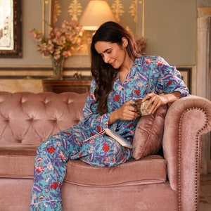 Peut inclure: Une femme portant un pyjama bleu et blanc &agrave; fleurs est assise sur un canap&eacute; en velours rose. Elle tient une tasse et un livre.