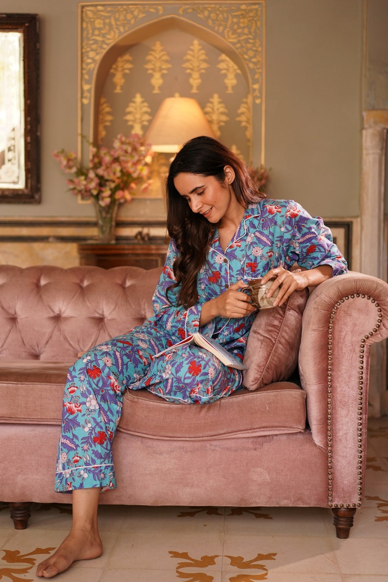 Peut inclure: Une femme portant un pyjama bleu et blanc &agrave; fleurs est assise sur un canap&eacute; en velours rose. Elle tient une tasse et un livre.