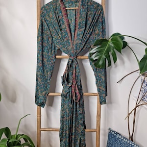 May include: A long-sleeved robe in a deep teal color featuring an intricate paisley pattern in shades of red, gold, and blue. The garment includes a matching fabric tie belt at the waist and a shawl collar with a contrasting thin red border trim.