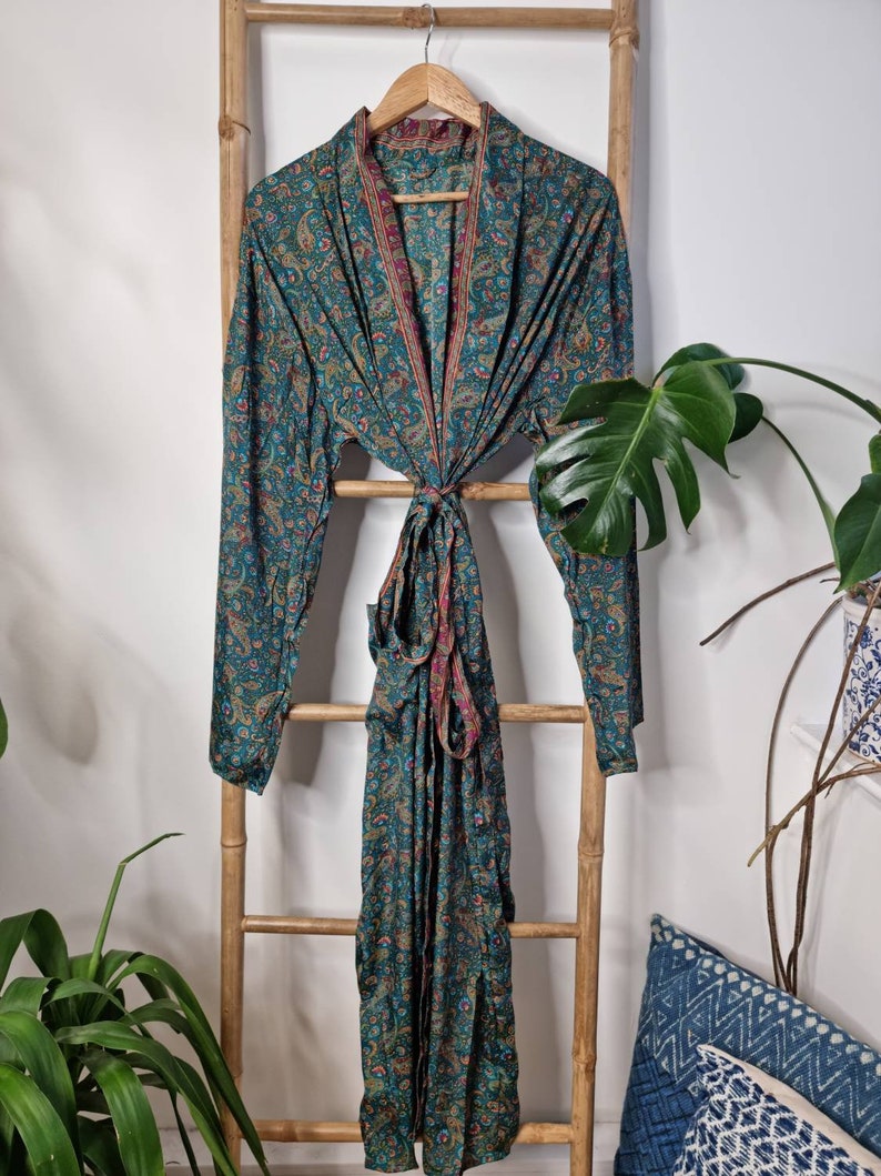 May include: A long-sleeved robe in a deep teal color featuring an intricate paisley pattern in shades of red, gold, and blue. The garment includes a matching fabric tie belt at the waist and a shawl collar with a contrasting thin red border trim.