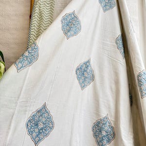 May include: White fabric with a repeating pattern of blue and white floral designs. The fabric is draped, showing the texture and flow of the material. The design is reminiscent of traditional block printing.