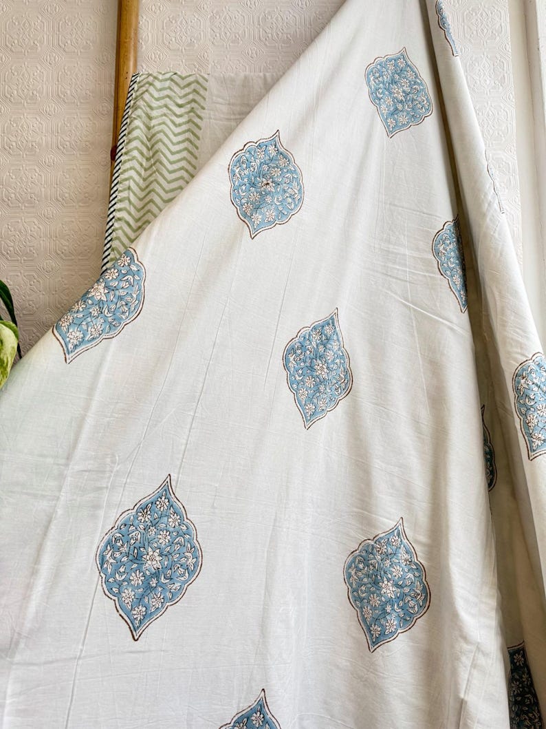 May include: White fabric with a repeating pattern of blue and white floral designs. The fabric is draped, showing the texture and flow of the material. The design is reminiscent of traditional block printing.