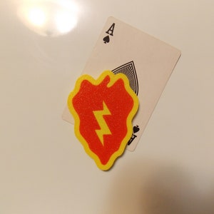 May include: A red and yellow felt patch with a lightning bolt design. The patch is shaped like a heart and has a jagged edge. The patch is sitting on top of an ace of spades playing card.