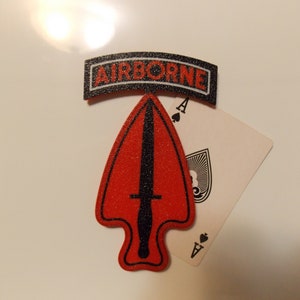 May include: A red and black military patch with the word "AIRBORNE" in white letters. The patch is shaped like a spearhead and has a black dagger in the center. The patch is sitting on top of an ace of spades playing card.