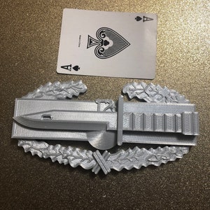 Puede incluir: Réplica gris impresa en 3D de una insignia militar, con una bayoneta, una corona de laurel y un clip de cartuchos. Un As de Picas está sobre la superficie. La insignia es detallada y parece ser de plástico.