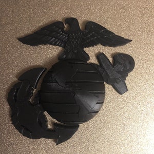 May include: A black 3D printed United States Marine Corps emblem. The emblem features an eagle perched atop a globe with an anchor.
