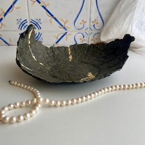 May include: A handcrafted, dark gray decorative bowl with gold leaf accents. The bowl has a textured surface and a unique, organic shape. A strand of white pearls is in the foreground. The bowl is likely used for home decor.