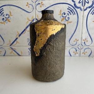 Pode incluir: Um vaso cilíndrico artesanal com uma superfície texturizada cinza escuro. Uma seção do vaso é adornada com folha de ouro. O vaso tem um pescoço preto e um design simples e elegante. Este vaso decorativo é adequado para decoração de casa.