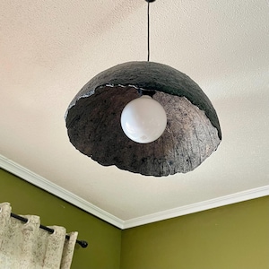 Puede incluir: Una lámpara colgante de color gris oscuro y texturizada con una gran pantalla redondeada. La pantalla tiene un aspecto rugoso y orgánico y rodea una bombilla esférica blanca brillante. La lámpara cuelga de un cable negro.
