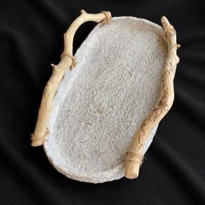 May include: An oval-shaped tray with a textured, off-white, stone-like base and two light brown, natural branch handles attached to the sides with thin, wrapped twine. The tray features a rough, uneven surface finish and organic, curved edges.