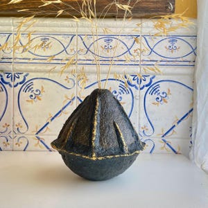 Puede incluir: Un jarrón gris oscuro, texturizado, con detalles dorados y una forma geométrica única. El jarrón está lleno de hierba seca de color dorado. El fondo presenta azulejos estampados con diseños azules y amarillos.