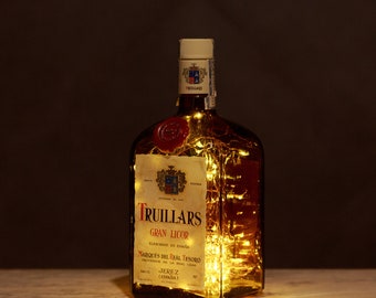 Op de afbeelding: Een doorzichtige glazen fles met een gouden etiket waarop "Truillars Gran Licor" en "Marqués del Real Tesoro" staat. De fles is gevuld met een gouden vloeistof en heeft een lichtsnoer erin.