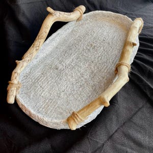 May include: Oval-shaped, light gray tray with a textured surface. Two wooden handles are attached with twine. The handles are light brown with a natural wood grain. The tray is on a black surface.