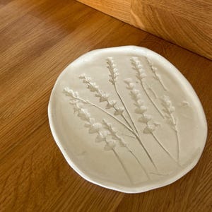 May include: Off-white ceramic plate with an embossed lavender design. The oval-shaped plate features a raised relief of lavender sprigs, creating a textured surface. The plate is set on a wooden surface.