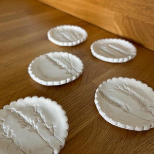 May include: Five white ceramic dishes with a scalloped edge, each featuring a raised wheat stalk design. The dishes are displayed on a wooden surface, highlighting their decorative and practical use. Each dish measures approximately 10 cm in diameter.