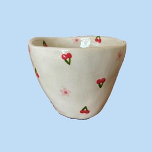 May include: A small, off-white ceramic bowl with an irregular, hand-formed rim. The exterior surface is decorated with a repeating pattern of small, bright red cherries with green stems and delicate, pale pink four-petalled flowers.