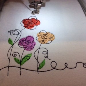 May include: Embroidered design of four stylized flowers in orange, purple, yellow, and green on a white fabric background.