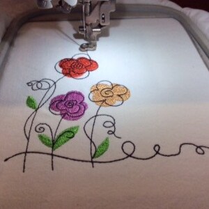 May include: Close-up of an embroidery machine stitching a design of four stylized flowers with black outlines and colored centers: red, orange, purple, and green.