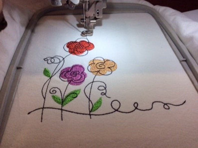 May include: Close-up of an embroidery machine stitching a design of four stylized flowers with black outlines and colored centers: red, orange, purple, and green.