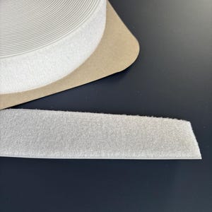 May include: A roll of white hook and loop tape, often called Velcro, with a strip of the tape extending out. The tape is white and has a soft, fuzzy texture. The roll sits on a brown cardboard backing.