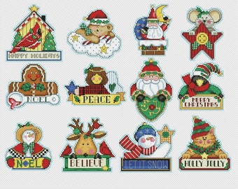 May include: Twelve colorful Christmas ornaments with various designs, including a gingerbread house, a snowman, a reindeer, a cat, and Santa Claus. The ornaments feature festive phrases like "Happy Holidays", "Noel", "Peace", and "Merry Christmas".