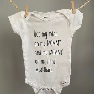 May include: White baby onesie with the text "Got my mind on my MOMMY and my MOMMY on my mind. #Laidback" printed in black. The onesie is hanging from a clothesline with wooden clothespins.