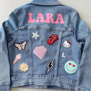 May include: A blue denim jacket with the name "LARA" in pink letters on the back. The jacket has various patches, including a butterfly, a diamond, a lightning bolt, a smiley face, a star, a tongue, a Hello Kitty, and an ice cream cone.