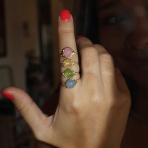May include: Four wire-wrapped rings with colorful, round gemstones are displayed on a finger. The gemstones are pink, yellow, green, and blue. The rings have a gold-colored wire setting.