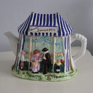 May include: A ceramic teapot shaped like a striped blue and white tent with the text Village Summer Fete. The exterior features a three-dimensional scene of people at a market stall with green grass, colourful figures, and blue streamers.