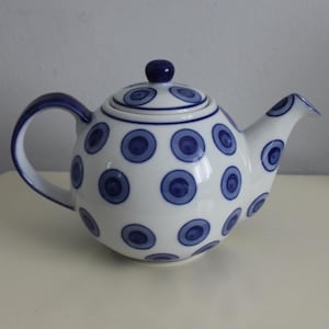 May include: White ceramic teapot with blue handle, spout, and lid. The teapot features a pattern of blue circles. The lid has a small, round blue knob. The teapot is on a white surface.