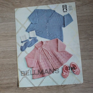 May include: A vintage pattern book featuring baby clothing. The cover displays a blue cardigan, pink cardigan, socks, and shoes. The cardigans have decorative cutouts. The text "BELLMANS" is visible on the cover.