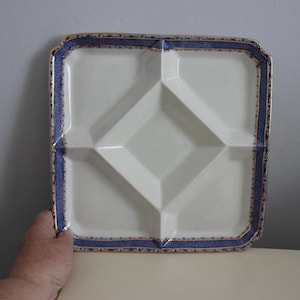 May include: Square white serving dish with four compartments and a diamond-shaped centre. The rim has a blue dotted pattern with gold trim. The dish is held by a hand.