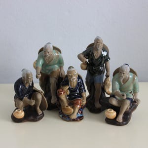 May include: Five ceramic figurines of elderly men in traditional clothing. The figures are in shades of blue, green, and brown, with detailed facial features and accessories. Each figure is approximately 10 cm tall.