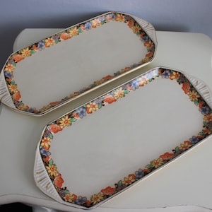 May include: Two rectangular serving platters with a floral border in orange, yellow, blue, and purple. The platters have a cream background and gold trim. They are on a white surface.