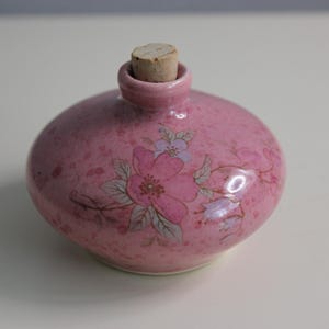 May include: A pink ceramic bottle with a cork stopper. The bottle has a speckled finish and is decorated with floral designs in pink, purple, and green. The bottle is round with a small neck.