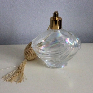 May include: A clear glass perfume bottle with an iridescent finish and a swirling, shell-like texture. The bottle features a polished gold-tone metal cap and is attached to a beige fabric-covered atomiser bulb with a matching beige tassel.