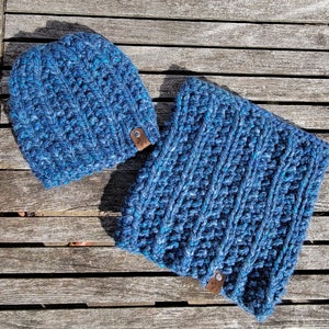 May include: A blue crocheted beanie and neck warmer set. The beanie has a brown leather tag with the word "Cozy" on it. The neck warmer is a long, rectangular piece of crocheted fabric.