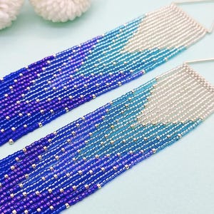Può includere: Un paio di lunghi orecchini a perline con un gradiente di perline blu, viola e argento. Gli orecchini sono realizzati con piccole perline rotonde e hanno un aspetto delicato e femminile.