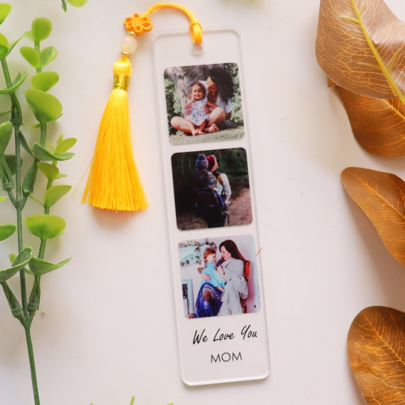 May include: A clear acrylic bookmark with three photo inserts and a yellow tassel. The photos show a mother and child. The bookmark has the text "We Love You MOM" printed on the bottom.