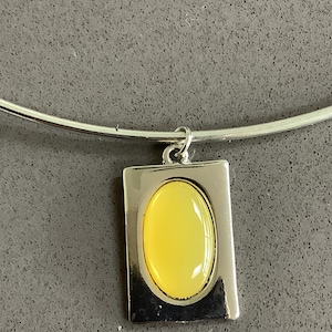 Puede incluir: Un collar de metal plateado con un colgante rectangular de acabado liso y pulido. Centrada en el rectángulo hay una piedra amarilla de forma ovalada y superficie lisa tipo cabujón. El colgante cuelga de un collar rígido de metal plateado.