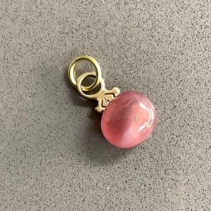 Peut inclure: Un charm doré avec une pierre précieuse rose de forme ovale. Le charm possède une petite boucle pour l'attacher à un collier ou un bracelet. La pierre précieuse est lisse et polie, d'aspect translucide.