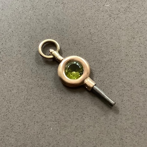 May include: A vintage watch key with a gold-tone setting and a green gemstone center. The key has a circular loop at the top and a dark metal stem. The key is likely used to wind a pocket watch.