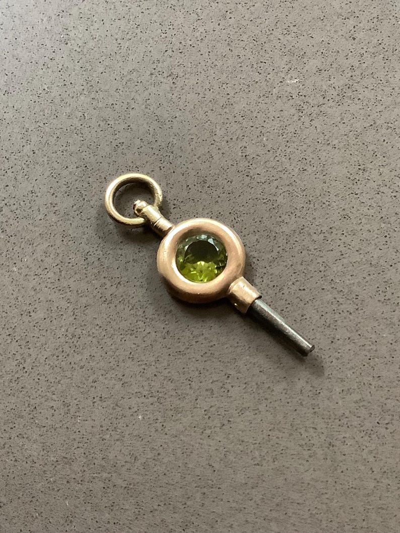 May include: A vintage watch key with a gold-tone setting and a green gemstone center. The key has a circular loop at the top and a dark metal stem. The key is likely used to wind a pocket watch.
