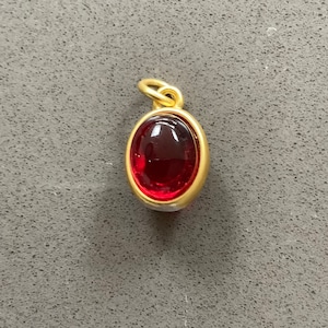 Puede incluir: Colgante de oro con una piedra preciosa roja oscura de forma ovalada. La piedra está engastada en un bisel de oro, con un pequeño lazo en la parte superior para una cadena. Una joya clásica.