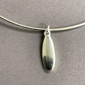 May include: A silver necklace featuring a teardrop pendant. The pendant is suspended from a thin, curved silver band. The background is a plain, light grey colour.