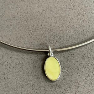 Könnte beinhalten: Eine silberfarbene Halskette mit einem hellgelben, ovalen Anhänger. Der Anhänger ist in einen silbernen Rahmen gefasst und hängt an einer dünnen, gebogenen Silberkette. Der Hintergrund ist einfarbig grau.