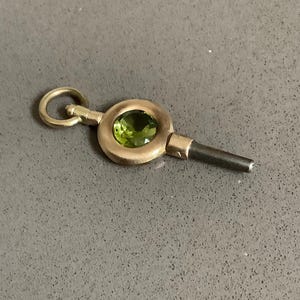 May include: A vintage gold-toned watch key with a circular design, featuring a vibrant green gemstone at its center. The key has a small loop at one end and a dark, cylindrical tip.
