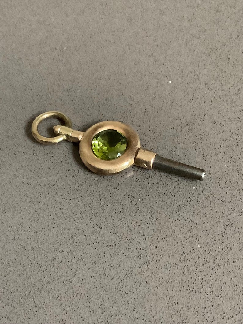 May include: A vintage gold-toned watch key with a circular design, featuring a vibrant green gemstone at its center. The key has a small loop at one end and a dark, cylindrical tip.
