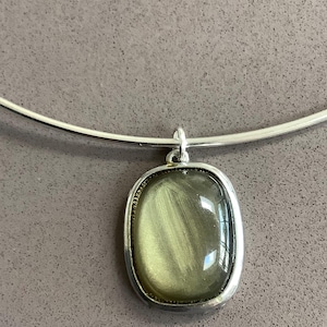Può includere: Una collana rigida in metallo color argento con un ciondolo rettangolare dagli angoli arrotondati. Il ciondolo contiene una pietra levigata color verde oliva chatoyant con una distinta striscia verticale riflettente al centro.